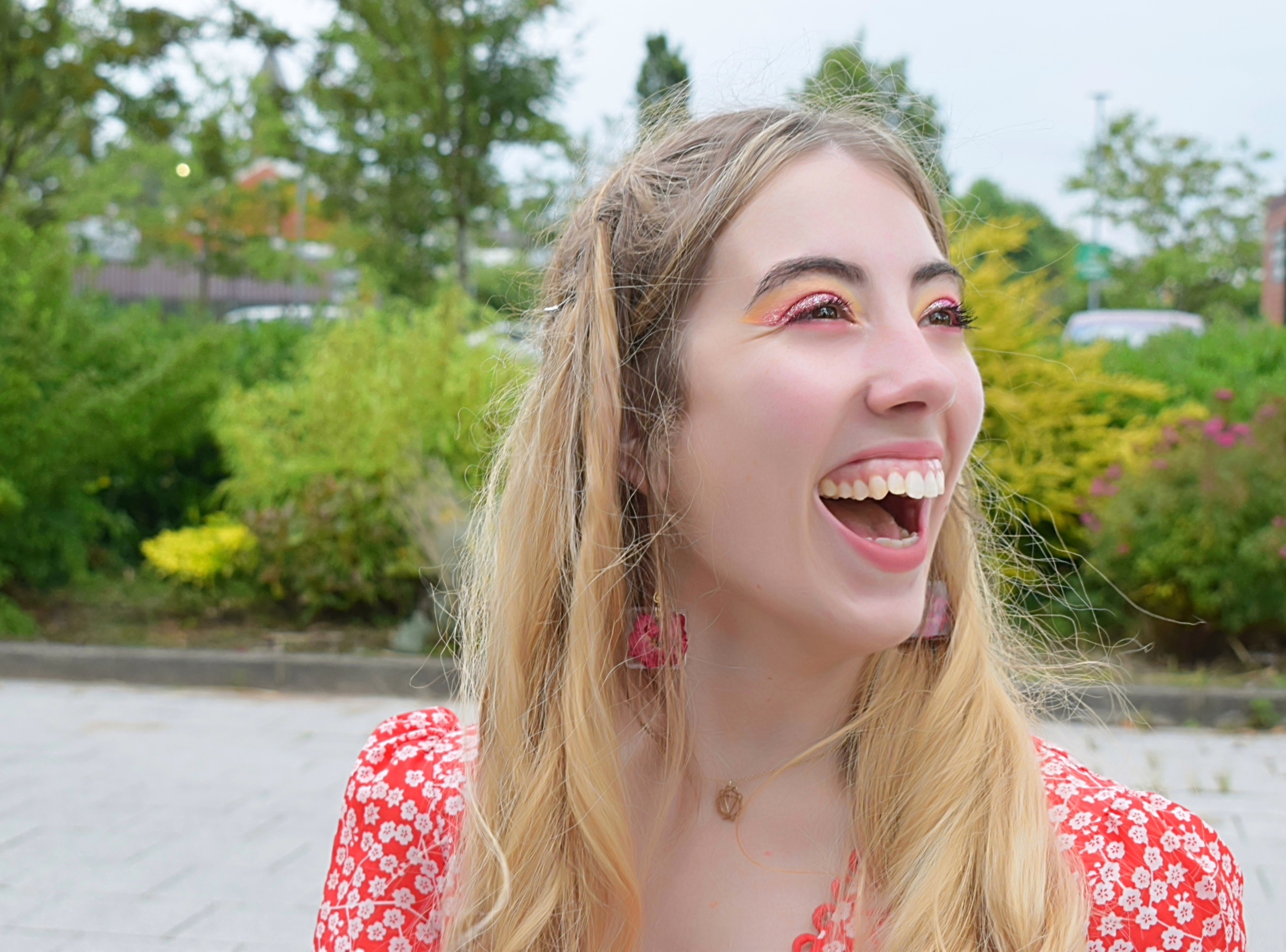 A young person in a leafy, outdoor setting with long blondish hair and brown eyes, wearing a  red dress and red flower earrings, smiling.